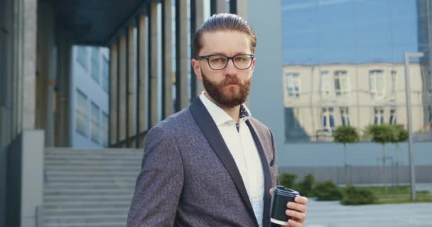 Snygg självsäker högkvalificerad 35-årig skäggig affärsman i formell kostym poserar på kamera med pappersmugg med kaffe nära modern kontorsbyggnad under paus — Stockvideo