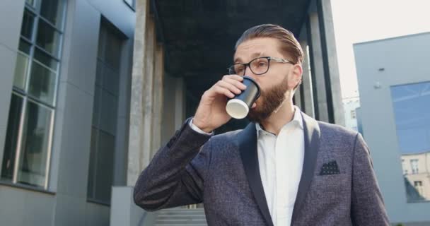 Vooraanzicht van positieve goed uitziende succesvolle lachende volwassen zakenman met goed verzorgde baard in glazen die koffie drinken in de buurt van modern stedelijk kantoorgebouw — Stockvideo