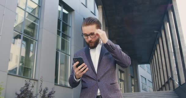 Primer plano de buen aspecto sorprendido exitoso adulto barbudo hombre de negocios en gafas que ver buenas noticias en la pantalla del teléfono, mientras que de pie cerca del centro de oficina en el descanso de tiempo — Vídeo de stock