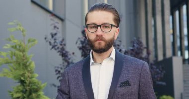 Gözlüklü ve şık takım elbiseli yakışıklı, kendine güvenen başarılı ofis müdürünün portresi. Gündüz vakti modern ofis binasının yanında poz veriyor.