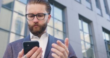 Çekici, ciddi, maksatlı, sakallı, modern ofis binasının yanında duran gözlüklü, akıllı telefonundan haber okuyan, ciddi bir ofis yöneticisi.
