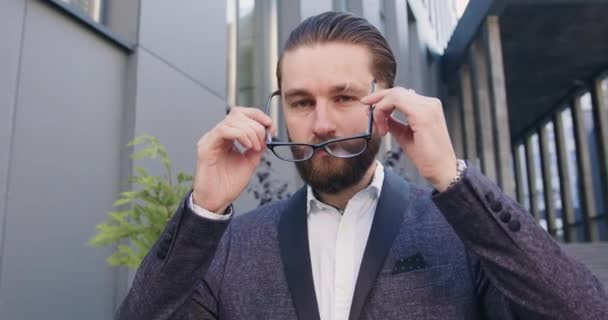 Close up of likable successful professional bearded man in business suit which putting on his glasses while loooking at camera with nice smile near modern office building — Stock Video