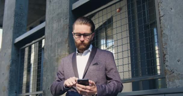 Close up de atraente homem barbudo adulto confiante concentrado em óculos e terno de negócios que espera por alguém enquanto usa seu telefone perto da parede do edifício moderno — Vídeo de Stock