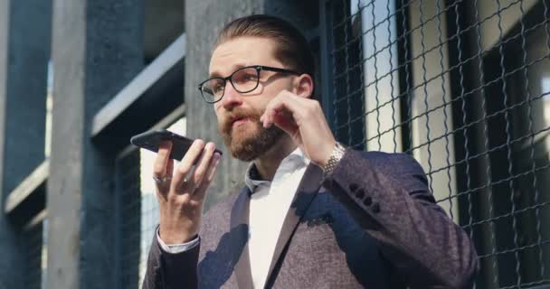 Guapo concentrado exitoso hombre de negocios barbudo con estilo en gafas está de acuerdo en reunirse con su socio de negocios por teléfono mientras está parado al aire libre cerca del edificio de oficinas — Vídeo de stock