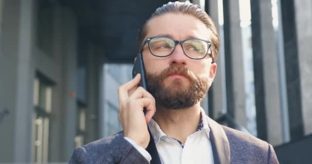 Primer plano de atractivo empresario barbudo positivo de 35 años de edad en gafas que habla en el móvil con su socio de negocios al aire libre en el fondo del edificio de oficinas — Vídeos de Stock