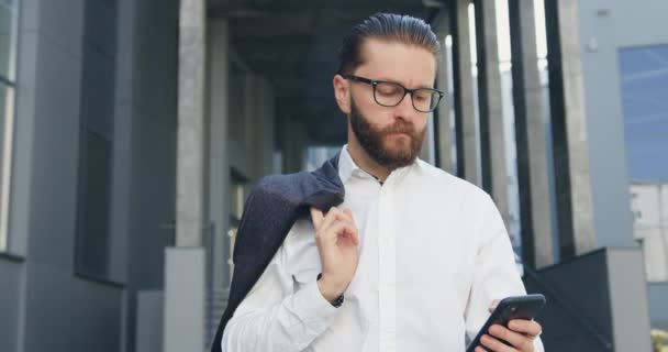 Primo piano di bello fiducioso responsabile ufficio adulto barbuto con camicia bianca che cammina vicino a un moderno edificio per uffici e utilizza il suo smartphone — Video Stock