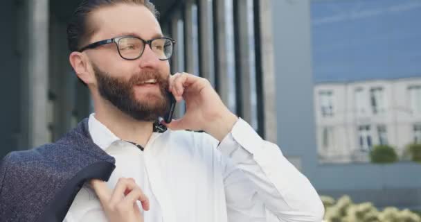 都会のビルの近くで休憩中に屋外で積極的な電話会話をしている眼鏡の35歳のひげそりのオフィスワーカーに満足した笑顔の可能性を閉じます — ストック動画
