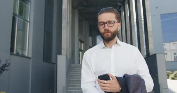 Portrait de beau concentré confiant 35 ans gestionnaire de bureau avec barbe bien entretenue dans des lunettes qui pose sur la caméra avec un visage sérieux tout en utilisant son mobile à l'extérieur près de l'entreprise, lent — Video