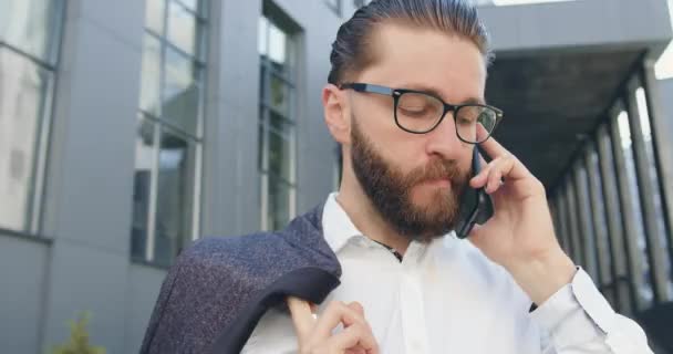 Close-up van goed uitziende geconcentreerde succesvolle doelgerichte bebaarde office manager in glazen die praten op de telefoon in de buurt van de stad modern gebouw — Stockvideo