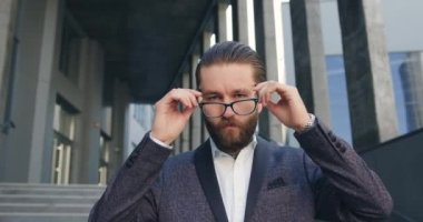 Güzel görünüşlü, kendine güvenen, gözlüklü, şık takım elbiseli yetişkin iş adamı ofis binasından çıkarken merdivenlerden aşağı iniyor ve kameraya bakıyor.