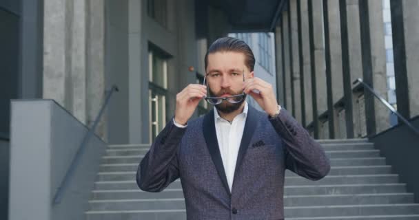 Vue de face d'attrayant homme d'affaires sérieux et confiant avec succès barbu en lunettes et costume élégant qui met des lunettes et pose sur la caméra avec les bras croisés près de l'immeuble de bureaux — Video