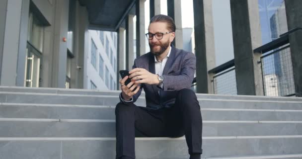 Handsome positive confident successful adult bearded office manager in glasses and business clothes sitting on steps and has video chat on smartphone during time break — Stock Video