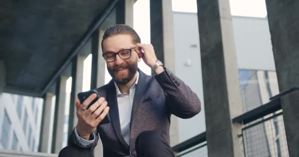 一位英俊的、面带微笑的、成功的成年大胡子办公室经理坐在城市建筑的台阶上，用他的手机浏览应用程序，4k — 图库视频影像