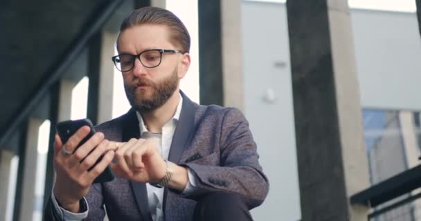 Bonito gerente de escritório barbudo sério e confiante em óculos sentados nos passos do prédio de escritórios e aplicativos de telefone de navegação controlando o tempo quando o intervalo de tempo terminar — Vídeo de Stock