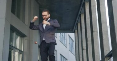 Yüksek moralli, başarılı, sakallı, gözlüklü, resmi kıyafetli yetişkin bir ofis çalışanı ofis binasından çıkarken dansla birlikte aşağı iniyor. Kapatın.