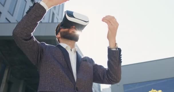 Handsome confident purposeful bearded businessman working on imaginary screen using augmented reality goggles standing on the background of modern urban building — Stock Video