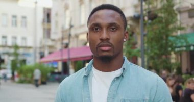 Yakışıklı, konsantre olmuş genç Afro-Amerikan modaya uygun gençlik kıyafetleri içinde eski şehirdeki sokak kafesinin yanında duruyor ve gündüz kameralarına bakıyor.