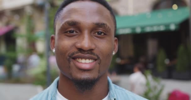 Beau dans la bonne humeur jeune afro-américain barbu souriant regardant la caméra avec un visage chanceux près d'un café de rue confortable le jour, au ralenti — Video