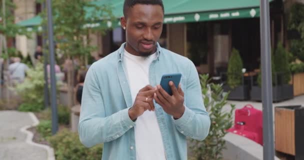Ймовірно, усміхнений сучасний молодий бородатий афроамериканець стоїть біля вуличного кафе і використовує свій смартфон для перегляду цікавих додатків, вид спереду — стокове відео