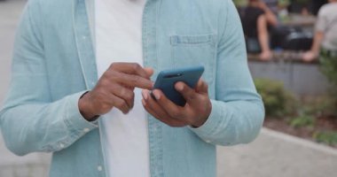 Bilinmeyen stil sahibi, siyah tenli, günlük kıyafetleri olan bir adam kafenin yanında duruyor ve telefon uygulamalarını tarıyor.