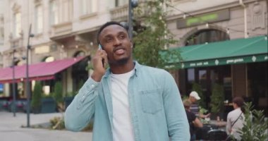 Güzel görünüşlü, gülümseyen, sıradan giysiler içindeki genç, siyah tenli adamın ön görüntüsü. Açık havada, sokak kafesi yakınlarında yaptığı telefon konuşmasından keyif alıyor.