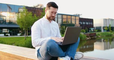 Modern cam bina arka planında çalışan serbest çalışan genç bir erkek. Çekici adam dışarıda dizüstü bilgisayarında sosyal medyayla sohbet ediyor. Yavaş çekim