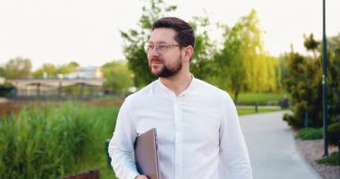 Dışarıda dizüstü bilgisayarlı, gülümseyen erkek çalışanın portresi. Genç girişimci şehir caddesi boyunca işyerine yürüyor. İş kariyeri. Yavaş çekim