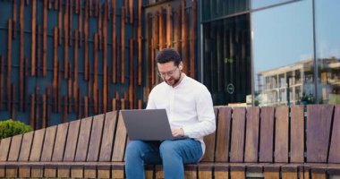 Beyaz gömlekli bir iş adamı bankta oturuyor ve modern cam binanın yanında dizüstü bilgisayarla çalışıyor. Odaklanmış genç adam uzaktan çalışıyor. Yavaş çekim