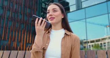 Elinde telefonla arkadaşıyla konuşan genç bir kadının portresi uzaktan kolay sesli mesaj, modern Ai teknolojisi konsepti, ses tanıma, modern bina arka planı üzerinden çevrimiçi çevirmen kullanıyor.