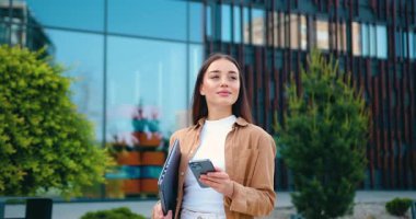 Güzel genç bir kadın, akıllı telefonuyla mesaj atarken ve dizüstü bilgisayarını tutarken ticari bir binanın önünden geçiyor. Şehir geçmişi. Modern yaşam tarzı, serbest çalışma ya da iş konsepti. Yavaş çekim