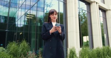 Kafkasyalı iş kadını resmi kıyafetlerle akıllı telefon kullanıyor, modern cam ofis binasının önünde mesaj yazıyor. Şehir merkezi, profesyonel yaşam tarzı, gündüz. Yavaş çekim