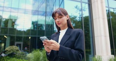 Açık havada, güneşli bir yaz gününde modern cam bir binanın önünde yürürken siyah ceketli gülümseyen esmer bir kadın akıllı telefon kullanıyor ve bir mesaj yazıyor. Yavaş çekim