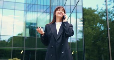 Resmi giysiler içinde gülümseyen genç bir iş kadını akıllı telefonuyla konuşuyor ve modern cam ofis binasının önünde dikilirken eliyle işaret ediyor. Dostça ve kendinden emin bir ifade. Şehir geçmişi. İletişim, kurumsal yaşam tarzı