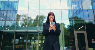 Resmi takım elbiseli, kendine güvenen genç ve gülümseyen bir iş kadını, iş çıkışı ve açık havada akıllı telefonunu kullanırken cam ofis binasından güvenle çıkıyor. Yavaş çekim