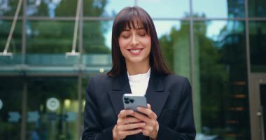 Gülümseyen bir iş kadınının resmi kıyafetleri içinde dışarıda yürürken ve akıllı telefon kullanırken. Ekrana tatmin olmuş bir ifadeyle bakıyor, arka plandaki ofis cam binasına karşı mesaj atıyor ya da olumlu haberler okuyor.