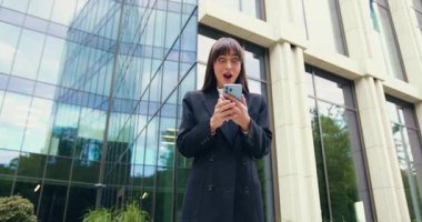 Şaşırtıcı iş kadınının elinde akıllı telefon var, bildirim aldığı için mutlu, ofisin arka planına karşı mutluluktan hareketler yapıyor, dışarıda. Duygular ve hoş duygular konsepti. Yavaş çekim