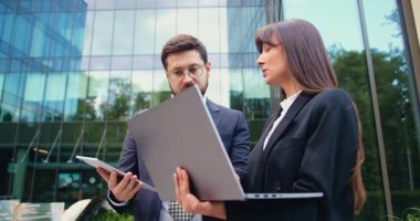 Açık havada ayakta duran iki iş adamı, modern bir şehir ortamında dizüstü bilgisayar ve tablet kullanarak uzaktan kumanda üzerinde çalışıyorlar. Yavaş motivasyon