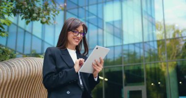 Resmi giyinmiş genç bir iş kadını modern bir ofis binasının önündeki bankta oturuyor, tablet üzerinde çalışıyor ve bir müşteriye yapay zeka sohbet robotu kullanarak mesaj yazıyor. Yavaş çekim