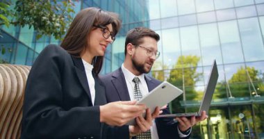 İki başarılı erkek ve kadının açık havada toplanıp, ofis binası yakınında dizüstü bilgisayar ve tablet kullanarak önemli iş konularını tartışmalarının yan görüntüsü. Yavaş çekim