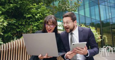 Modern cam ofis binasının dışındaki bankta oturmuş, büyük bir iş başarısını kutlamak için dizüstü bilgisayar ekranına bakarken heyecanla gülümseyen ve beşlik çakan neşeli iş kadını ve iş kadınının yan görüntüsü. Yavaş çekim