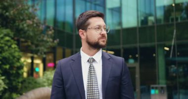 Ciddi ve başarılı, takım elbiseli ve gözlüklü beyaz bir işadamının yakın plan portresi modern bir ofis binasının önünde dikilip, güvenle kameraya bakıyor, bir şirket yöneticisinin vesikalık resmi. Yavaş çekim