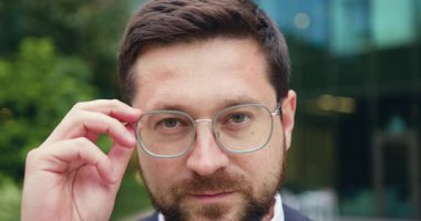 Gözlüklerini ayarlayan gülümseyen beyaz bir adamın yakın plan çekimleri ve dışarıda bulanık bir arka plan ile kameraya bakması. Yavaş çekim