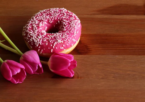 Donut con glaseado rosa y tulipanes rojos sobre un fondo de madera oscura. Vista superior. Concepto alimenticio . — Foto de Stock