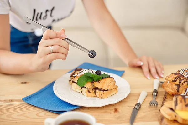 Female hands with kitchen tongs decorating sweet croissant on cooking table