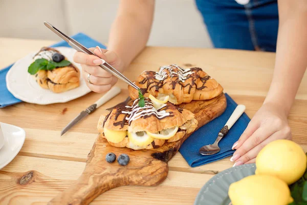 Female hands with kitchen tongs decorating sweet croissants on cooking table