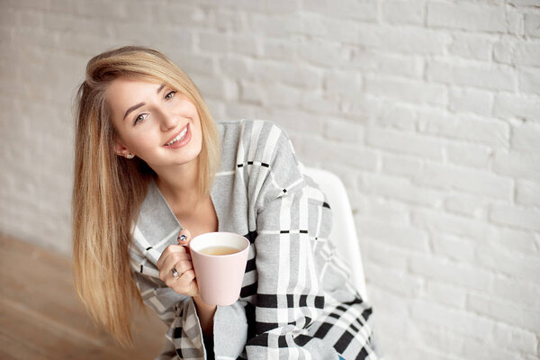 Young happy girl wrapped in a warm blanket looks happy holding cup of tea indoors