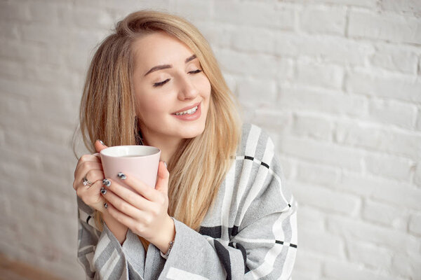 Young happy girl wrapped in a warm blanket looks happy holding cup of tea indoors