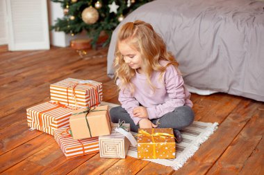 Noel sabahı bir kız pijama sarı saçları ile yatağın oturan ve hediyeler Noel ağacı arka planda bakarak. Yeni yıl, Noel kavramı.