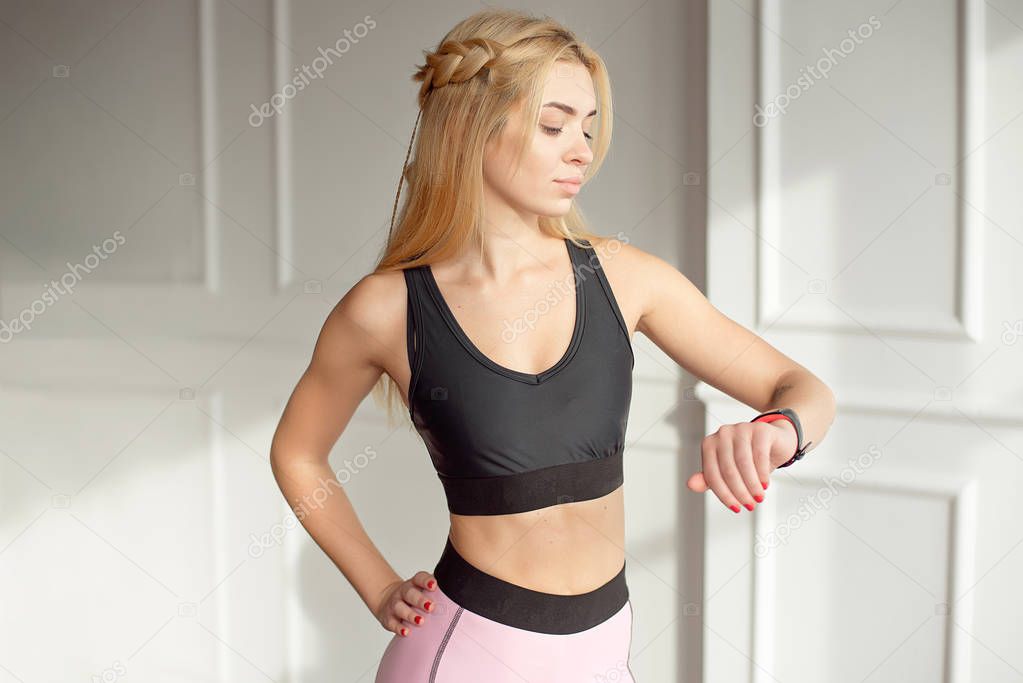 A young slim woman with an athletic body with long blond hair, wearing a black top and pink leggings in sportswear, is standing in a bright yoga room with a large panoramic window, looking at her