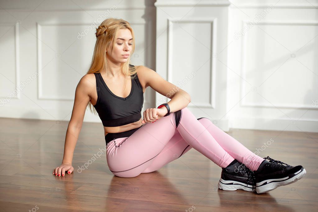 Young slim woman with an athletic body with long blond hair, dressed in black sportswear and pink leggings, sits on the floor in a bright yoga room with a large panaramic window, looks at her fitness